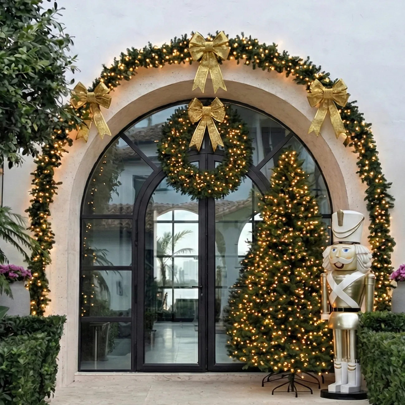 Arched glass entrance framed by a lit garland and gold bows, with a wreath, decorated Christmas tree, and gold nutcracker.