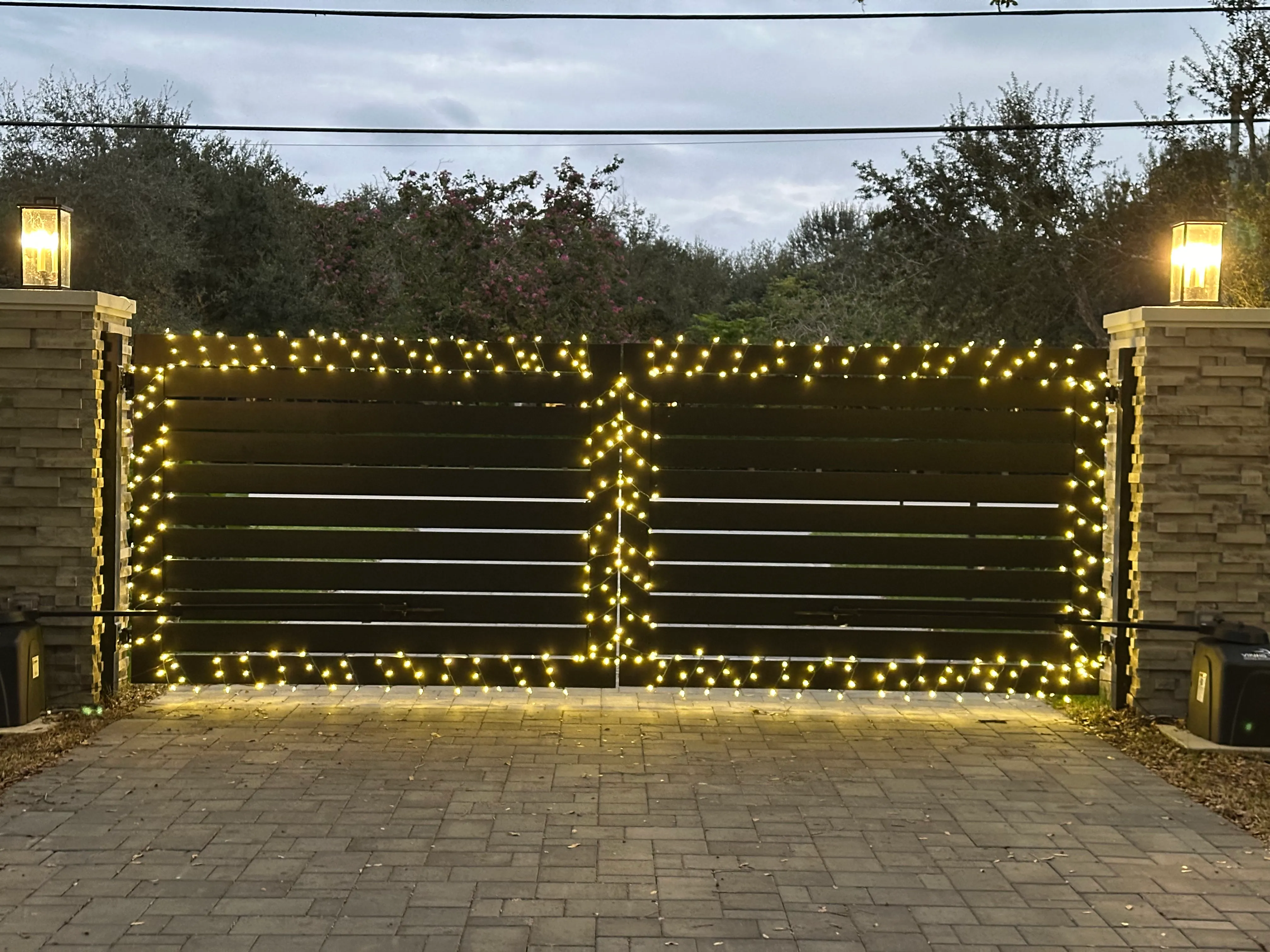 Dark metal entrance gate outlined with warm white Christmas lights between stone pillars.