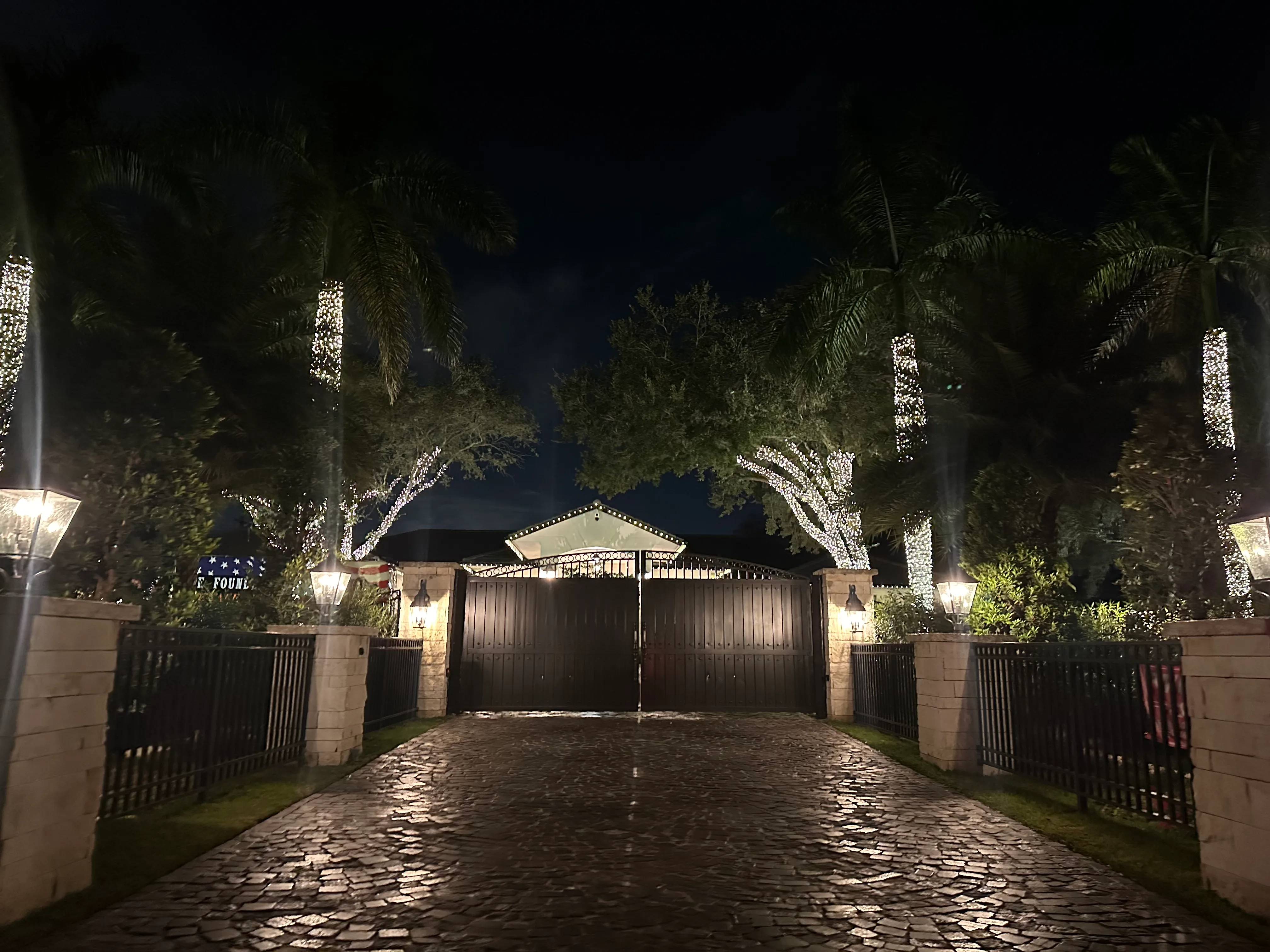 Gated stone driveway framed by palms and trees wrapped in warm white Christmas lights, with an illuminated home beyond the gate.